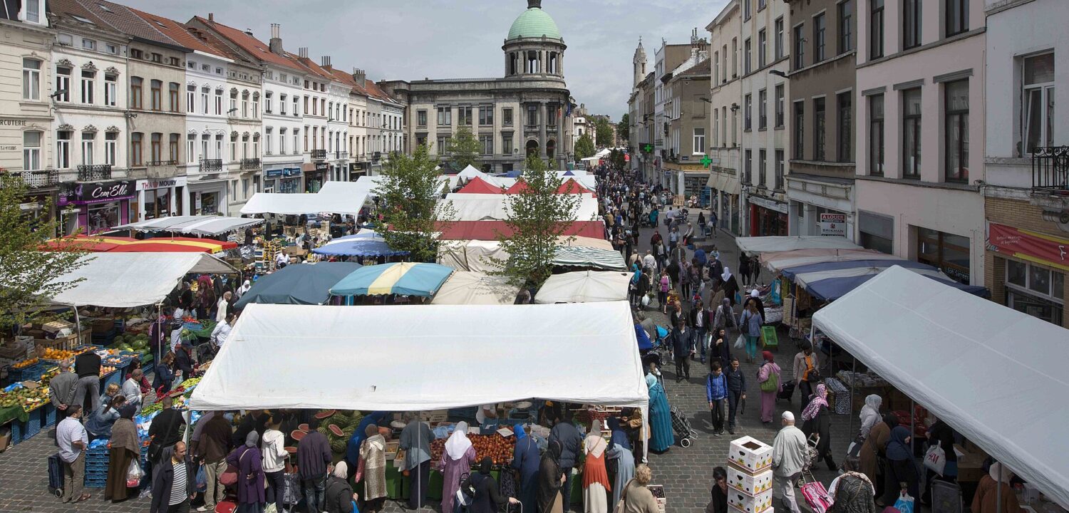 Crowded open-air food market seen from above in Brussels' Molenbeek district, with stalls of fruit and vegetables, shoppers with trolleys and a green-domed building at the end of the street