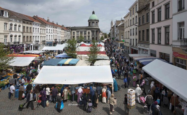 Crowded open-air food market seen from above in Brussels' Molenbeek district, with stalls of fruit and vegetables, shoppers with trolleys and a green-domed building at the end of the street