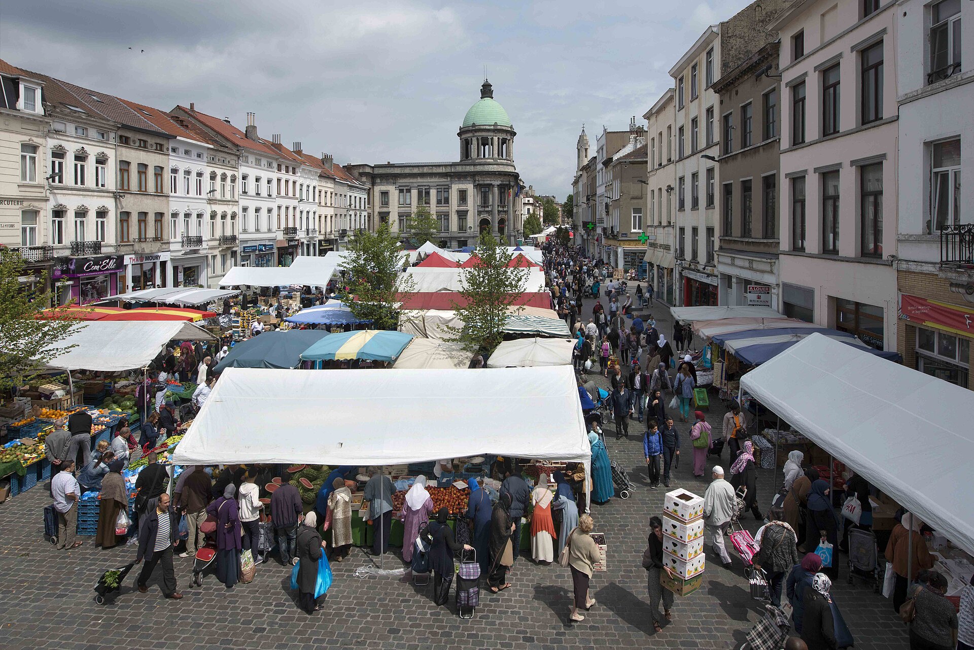 Crowded open-air food market seen from above in Brussels' Molenbeek district, with stalls of fruit and vegetables, shoppers with trolleys and a green-domed building at the end of the street