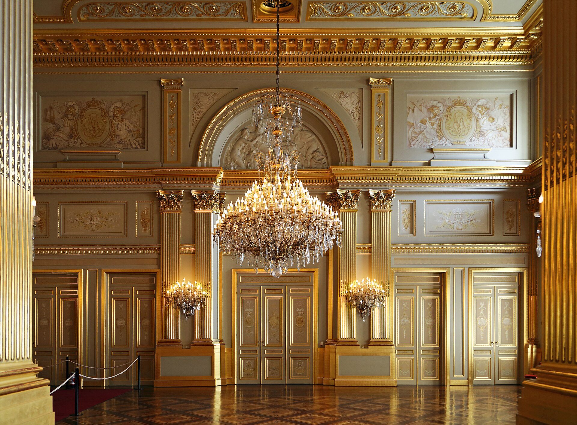 A crystal chandelier hangs between gilded pilasters in the empty Throne Room of the Royal Palace of Brussels, above polished parquet flooring.