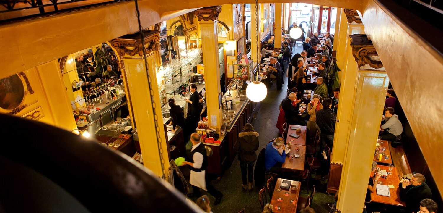Customers drink at wooden tables beneath globe lamps inside Brussels A la Mort Subite, seen from the upper gallery.