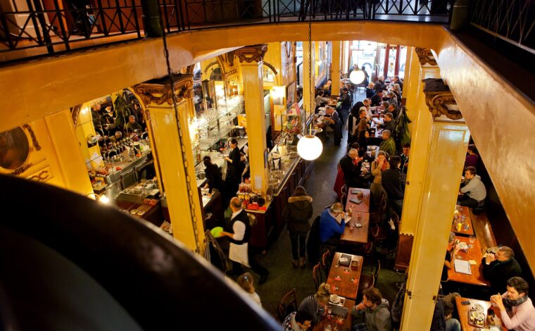 Customers drink at wooden tables beneath globe lamps inside Brussels A la Mort Subite, seen from the upper gallery.
