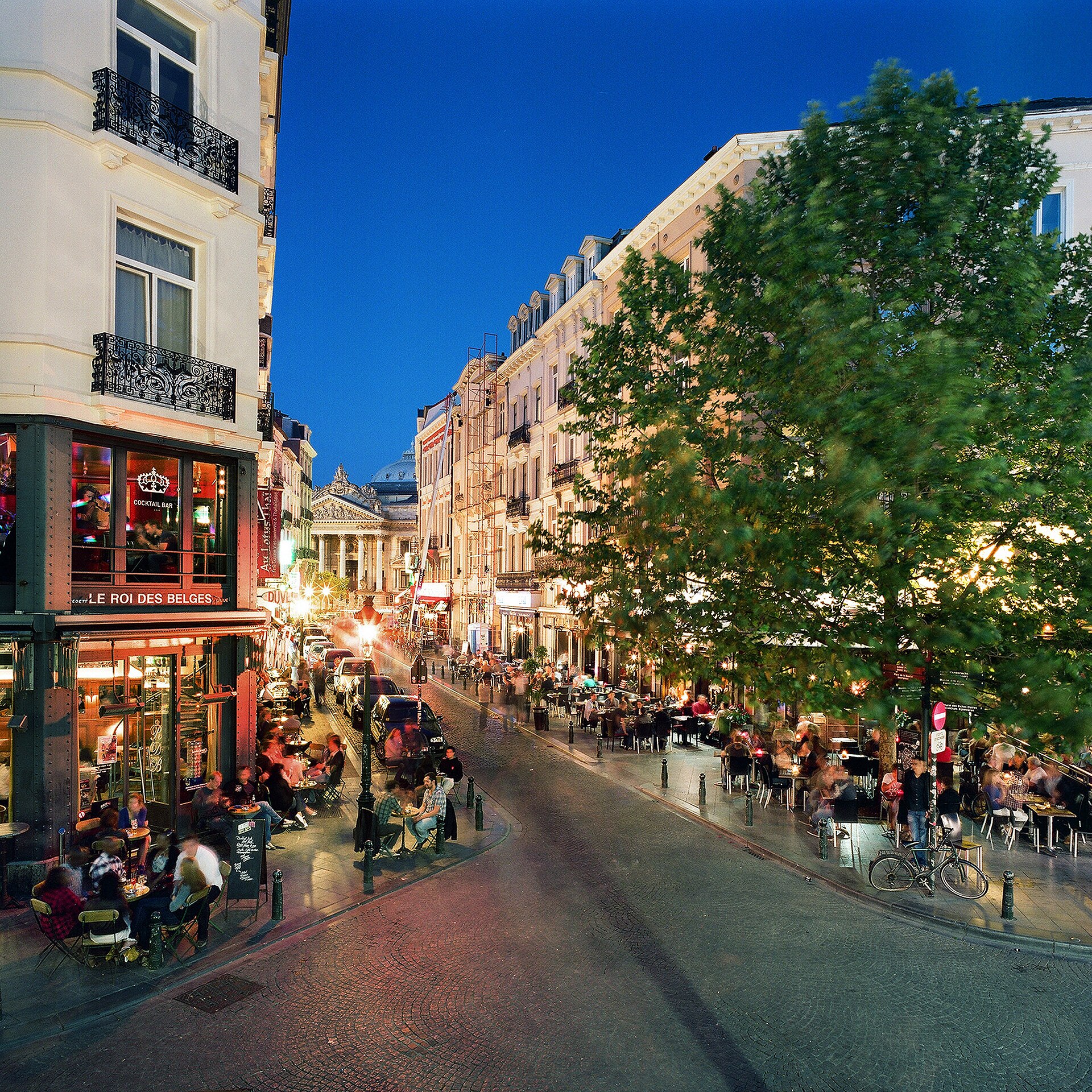 Customers fill cafe terraces beside Le Roi des Belges on Place Saint-Gery in Brussels beneath a deep blue evening sky.