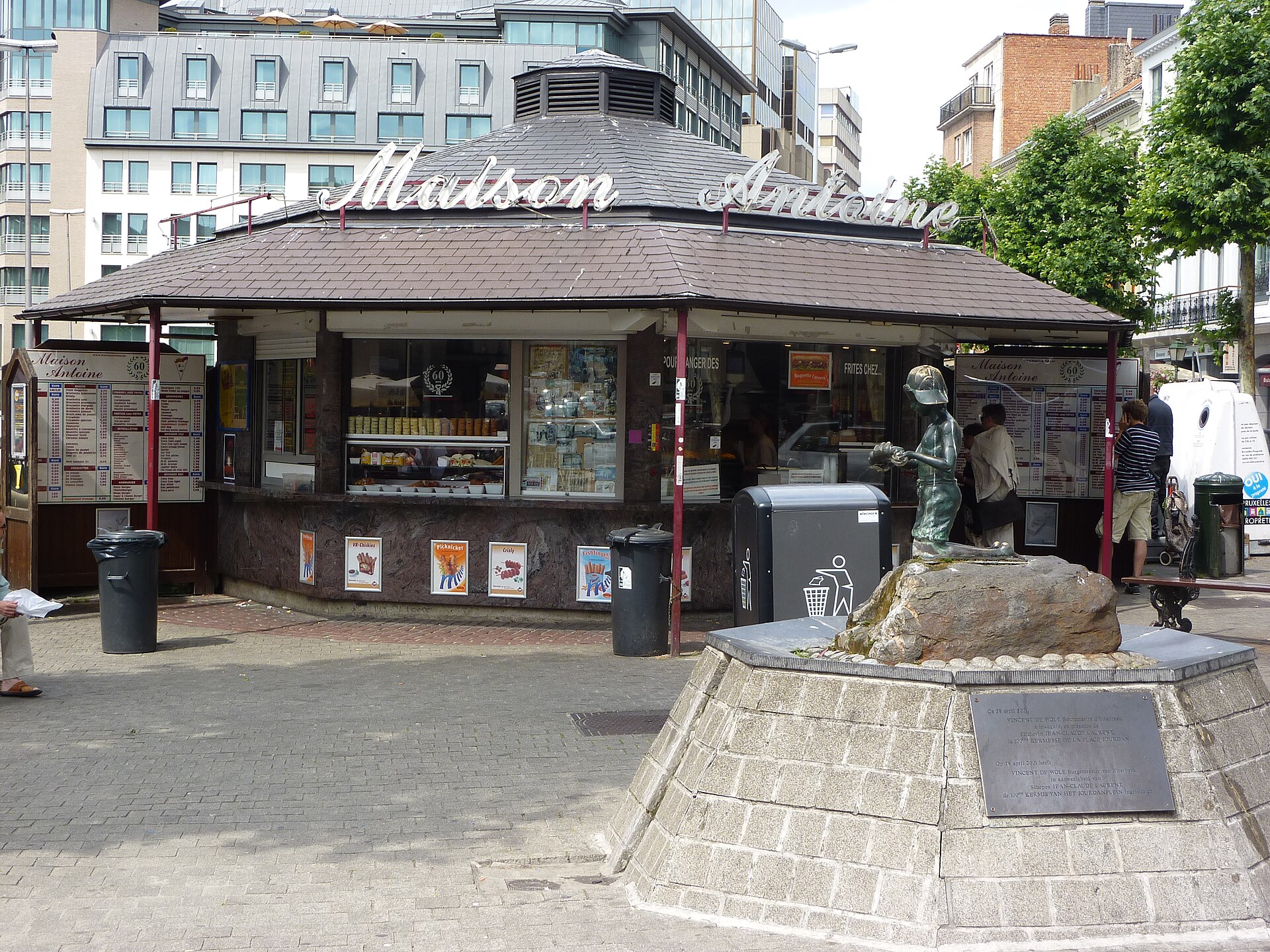 Customers order at the Maison Antoine frites kiosk on Place Jourdan in Brussels, behind a small fountain with a bronze figure.