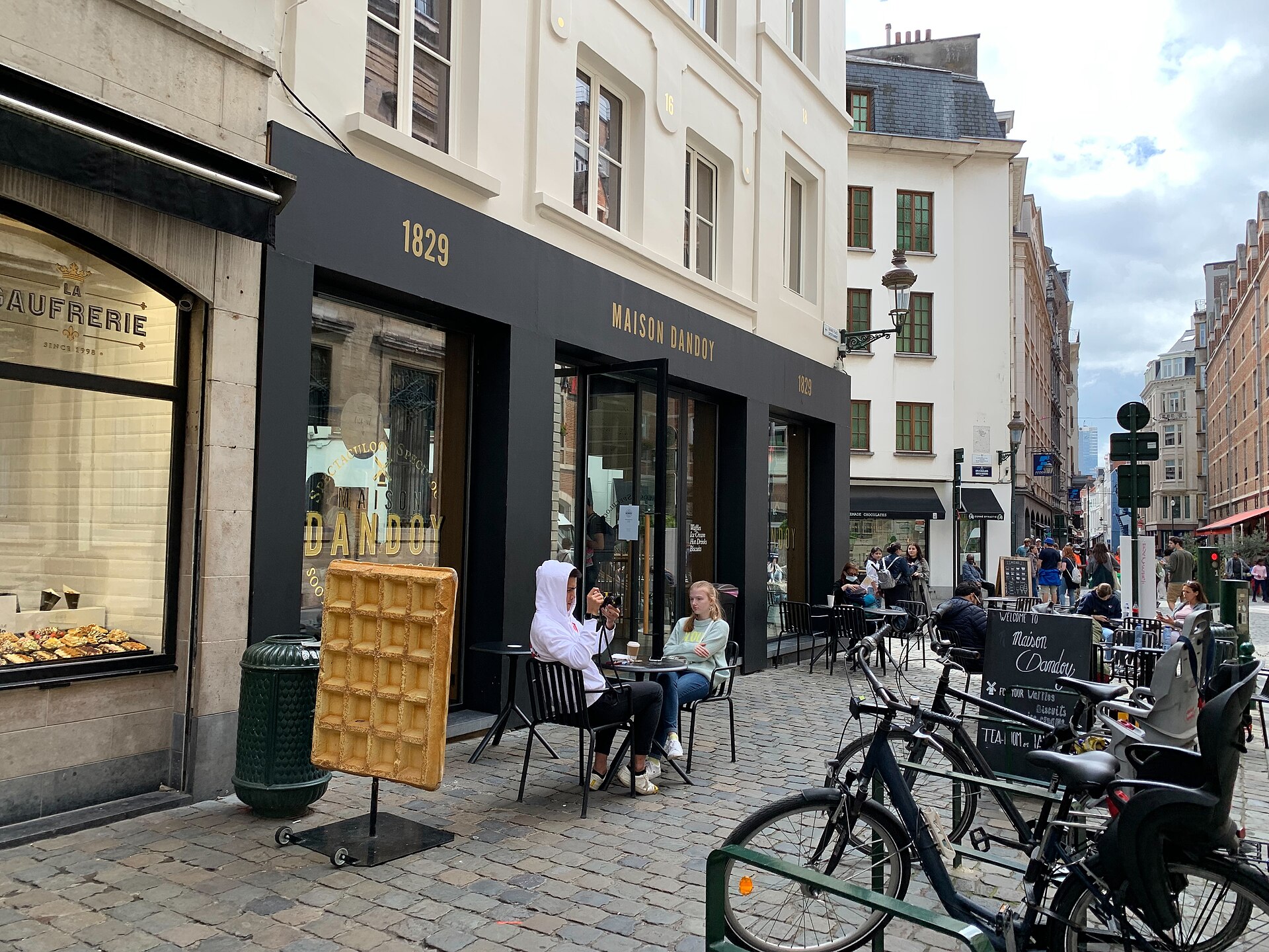 Customers sit outside Maison Dandoy in Brussels beside a giant waffle model and a neighbouring window displaying topped waffles.