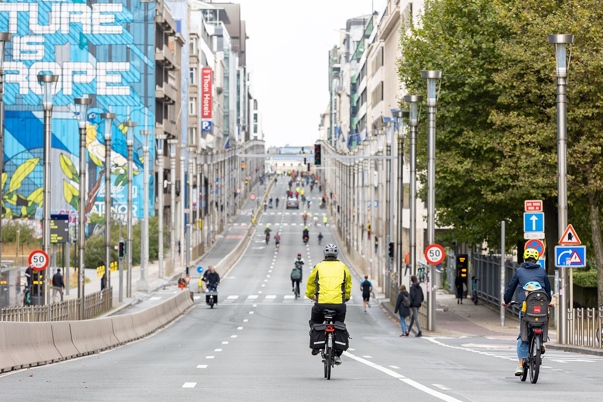 Cyclists ride along a broad Brussels street on Car Free Day, passing office buildings and a large blue mural.