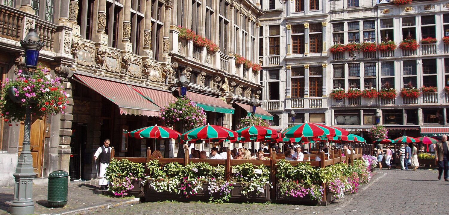 Diners sit beneath red and green parasols on a restaurant terrace framed by ornate guildhall facades on the Grand-Place in Brussels.