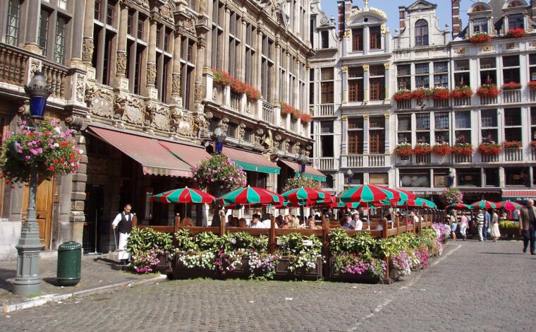 Diners sit beneath red and green parasols on a restaurant terrace framed by ornate guildhall facades on the Grand-Place in Brussels.