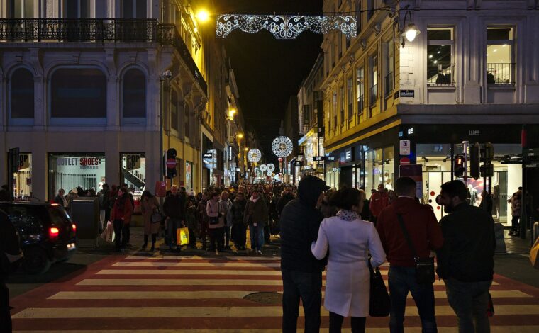 Evening shoppers crowd Rue Neuve in Brussels beneath decorative lights, with illuminated shop windows lining both sides.