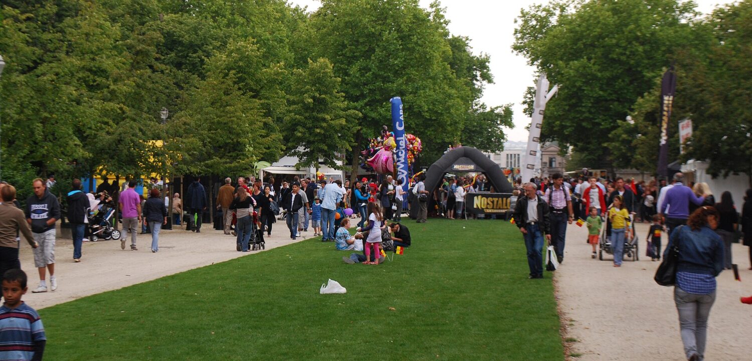 Families walk beside lawns and festival stalls in Brussels Park, with balloons and strollers among the National Day crowds.