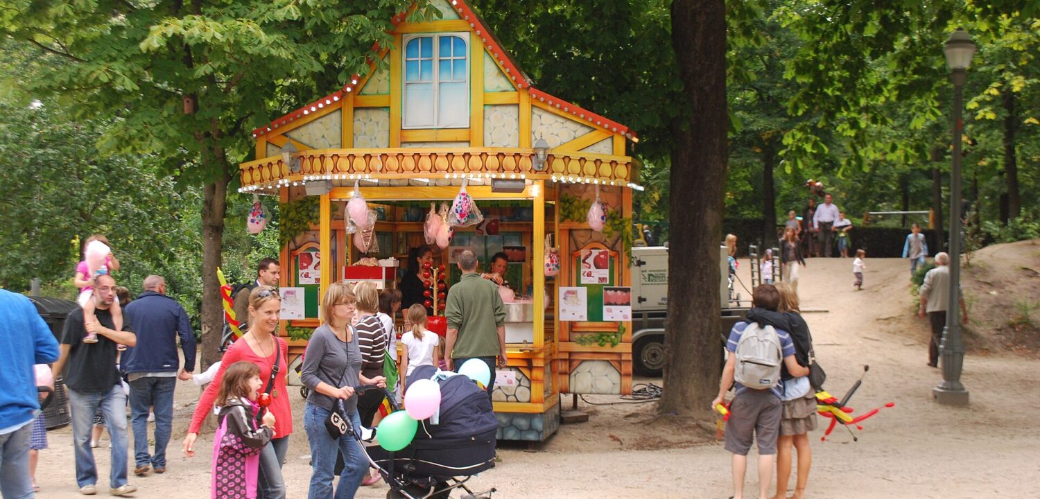 A family with a stroller passes a colourful sweet stall beneath leafy trees in Brussels Park during National Day celebrations.