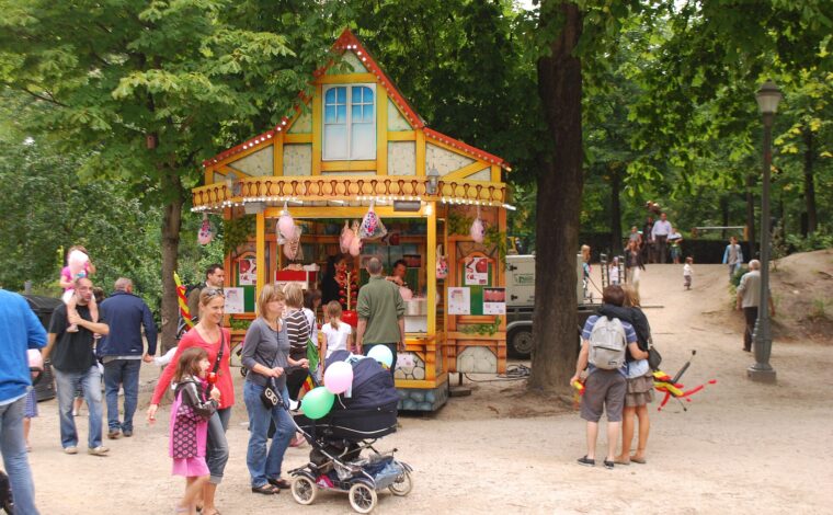 A family with a stroller passes a colourful sweet stall beneath leafy trees in Brussels Park during National Day celebrations.