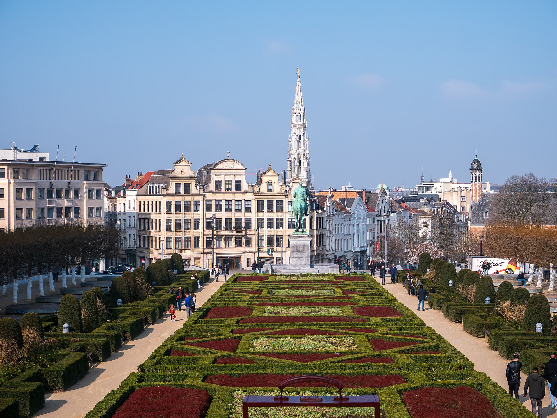 Geometric flower beds frame walkers in the Mont des Arts garden, Brussels, beneath the Albert I statue and Town Hall spire.