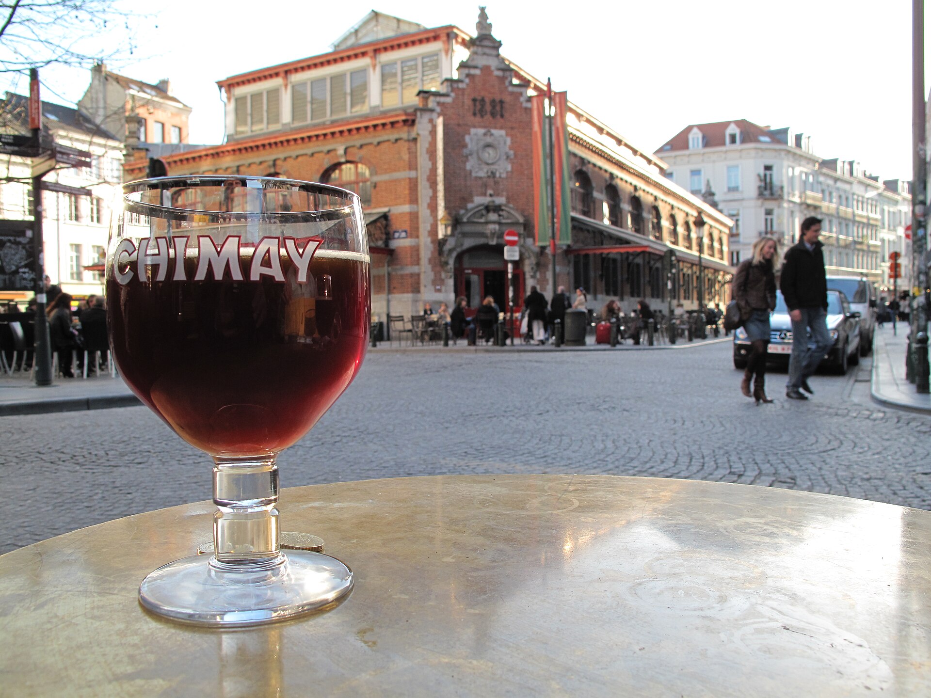 A glass of Chimay beer stands on a Brussels cafe table facing the red brick Halles Saint-Gery and passing pedestrians.