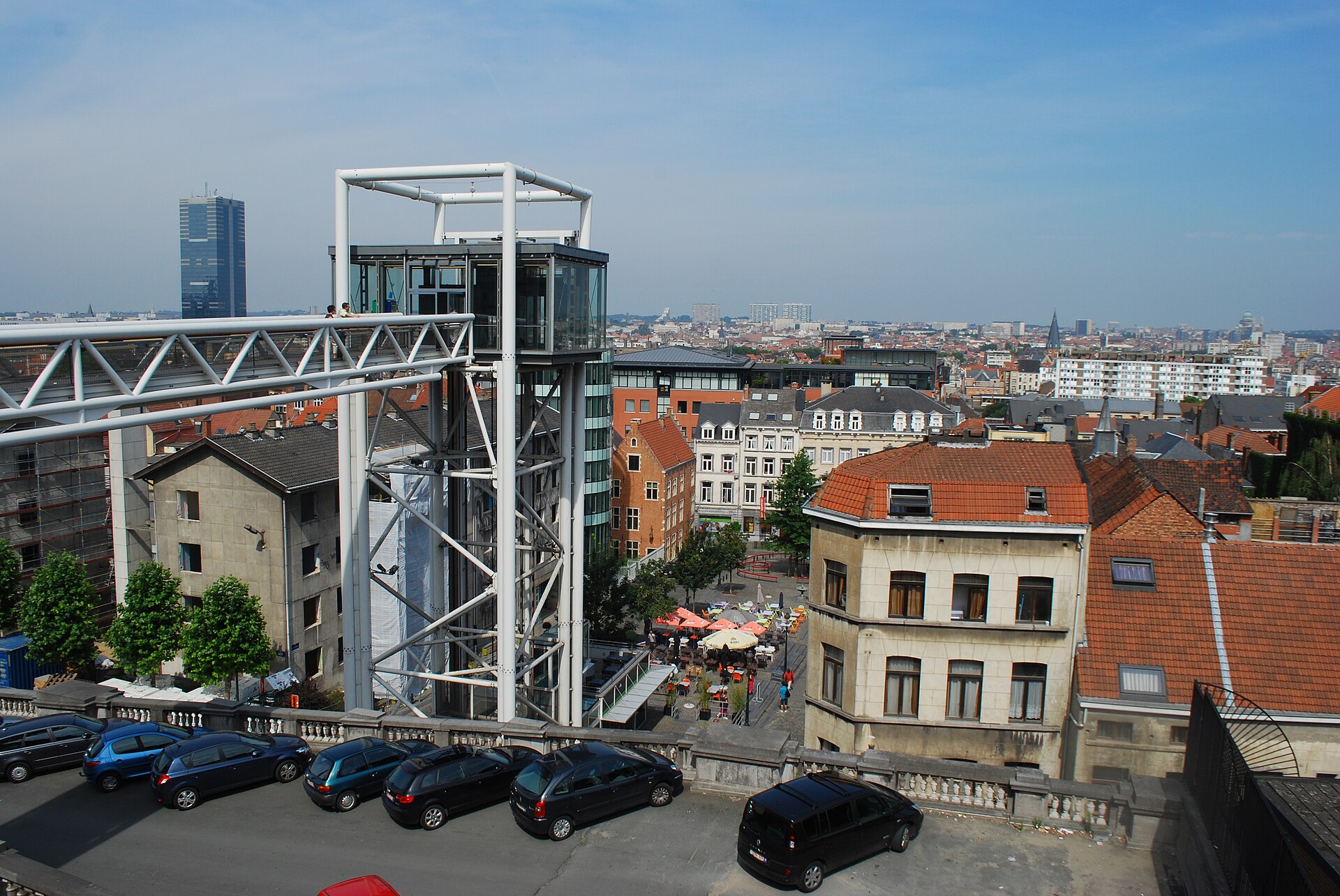 The glass Marolles lift and steel footbridge rise above red rooftops in Brussels, with people crossing the bridge.