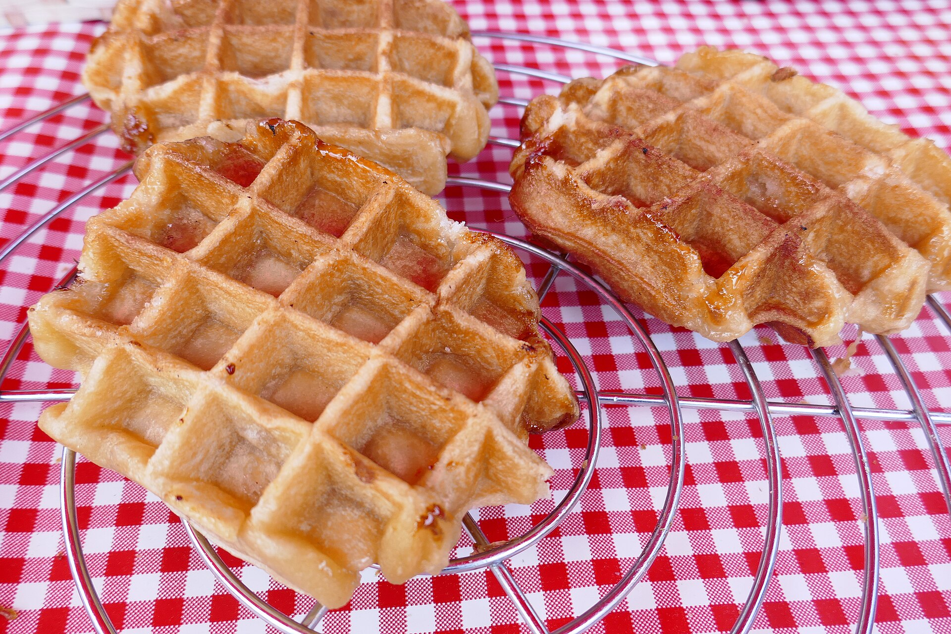 Golden Liege waffles with irregular edges and caramelised sugar rest on wire racks above a red and white checked tablecloth.