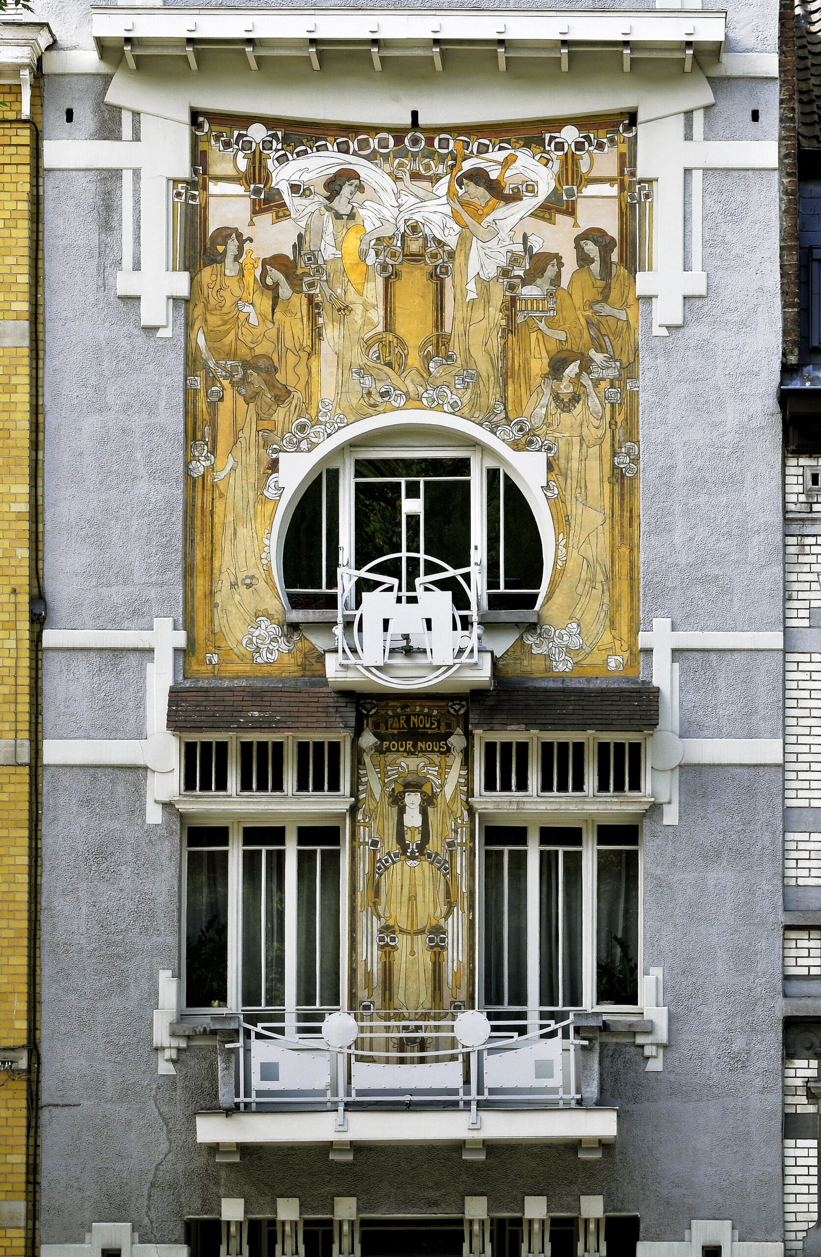 Golden sgraffito women surround a circular window and white balcony on the Maison Cauchie facade in Etterbeek, Brussels.