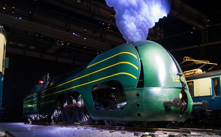Green locomotive 12.004 stands beneath a decorative smoke cloud inside Brussels’ Train World museum, with another historic train behind.