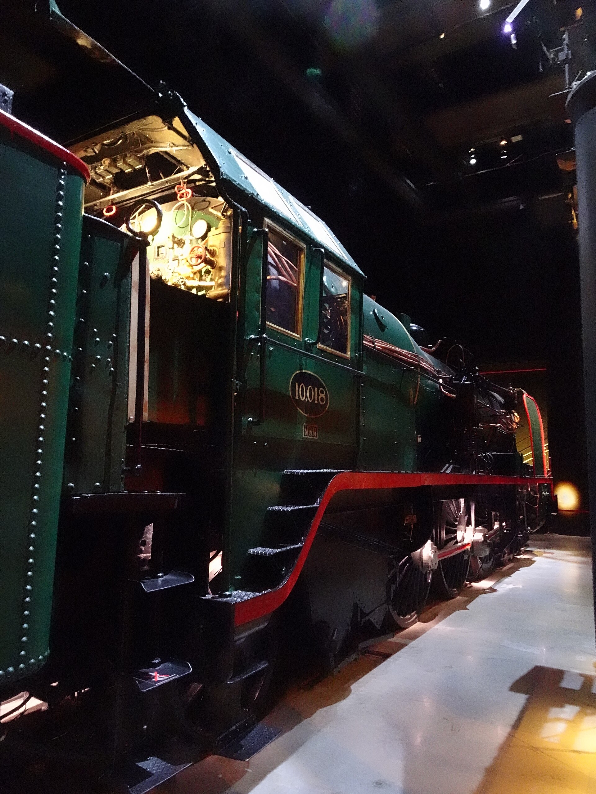 Green steam locomotive 10.018 stands inside Train World in Brussels, with illuminated cab controls, red trim and large wheels.