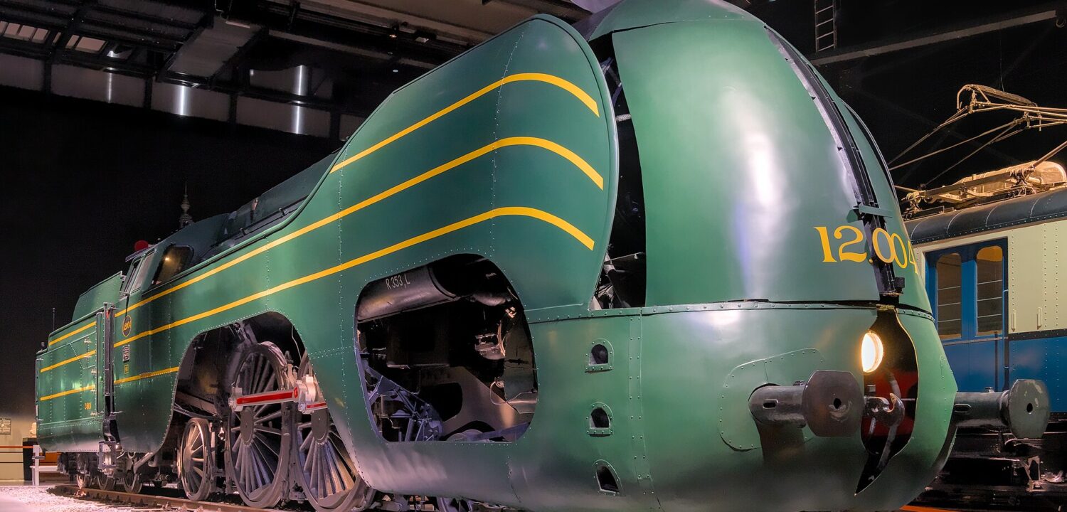 Green streamlined locomotive 12.004 stands inside Train World in Brussels, with yellow stripes, exposed driving wheels and a glowing headlamp.