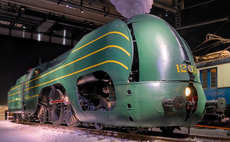 Green streamlined locomotive 12.004 stands inside Train World in Brussels, with yellow stripes, exposed driving wheels and a glowing headlamp.