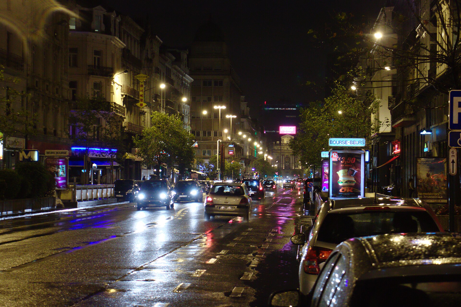 Headlights and street lamps reflect on wet Boulevard Anspach in Brussels beside the illuminated Bourse-Beurs station sign at night.