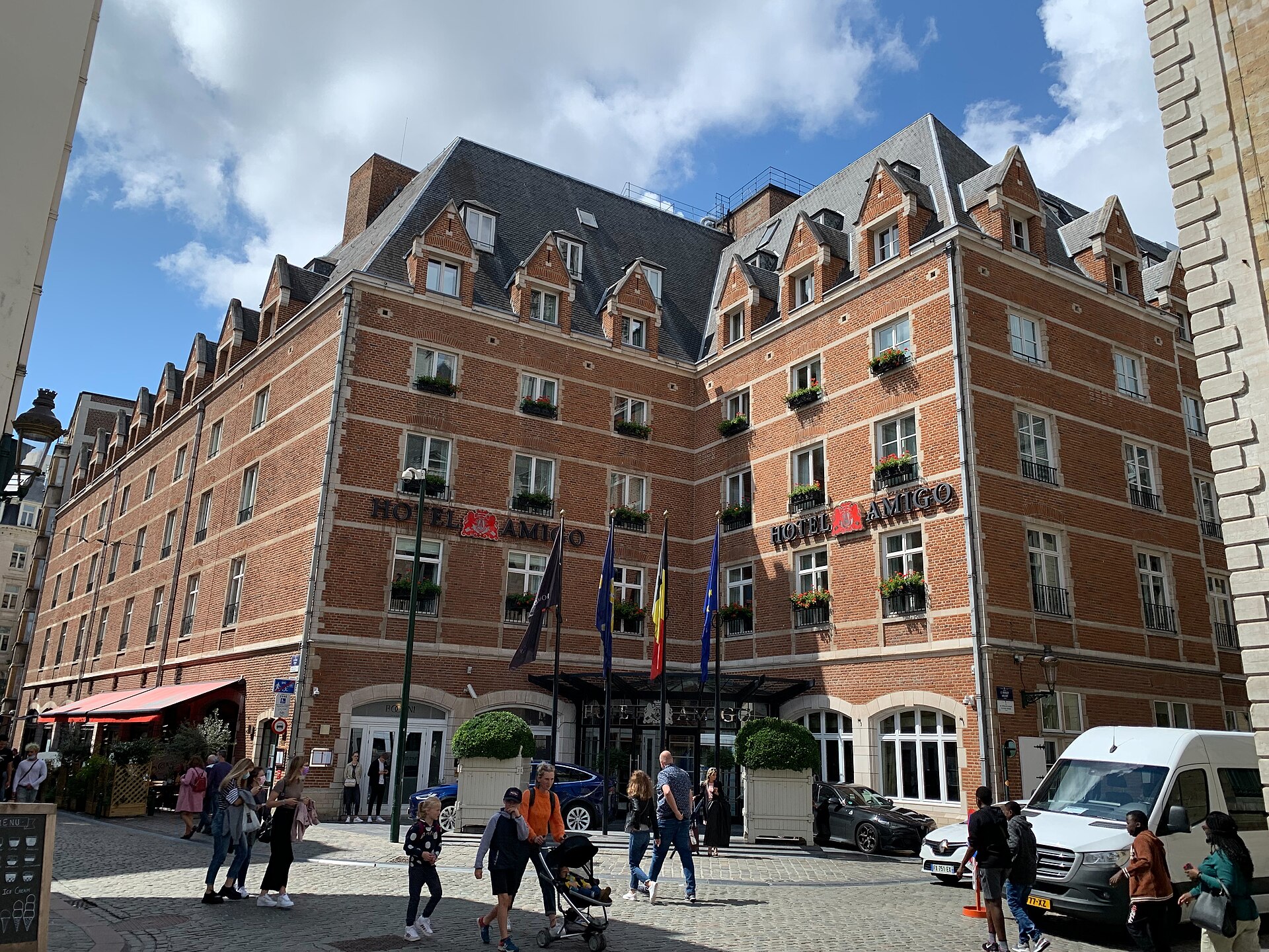 Hotel Amigo’s red-brick facade in Brussels rises above flags and flower boxes, with pedestrians and a stroller crossing the cobbled street.