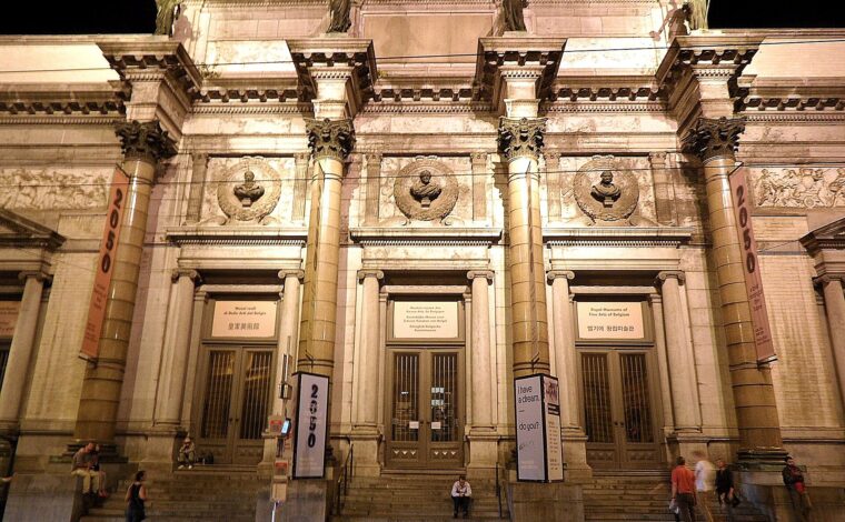 Illuminated columns and bronze rooftop statues at the Royal Museums of Fine Arts of Belgium in Brussels, with people outside after dark.