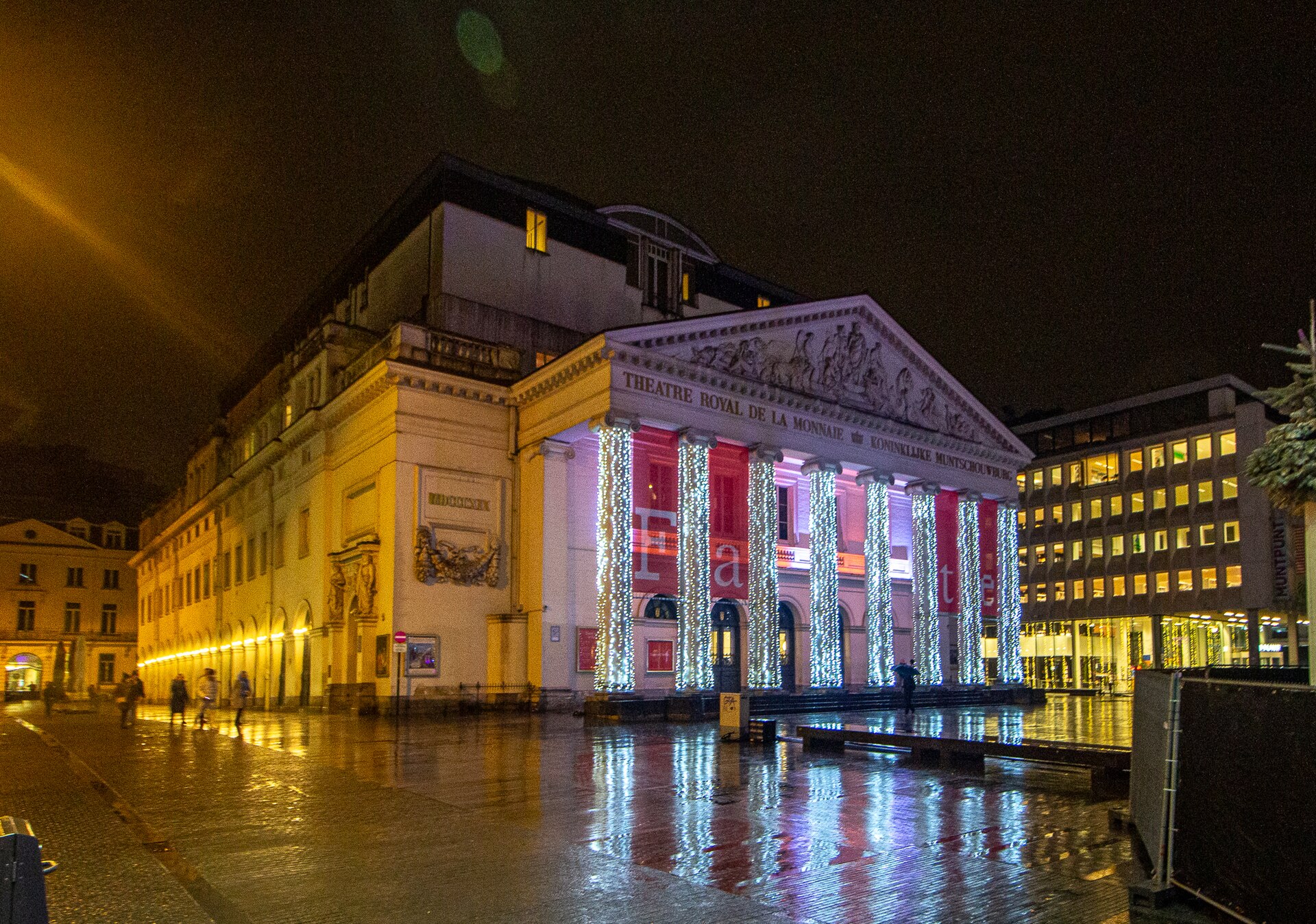 Illuminated columns of La Monnaie opera house reflect across the wet square in Brussels on a December evening.