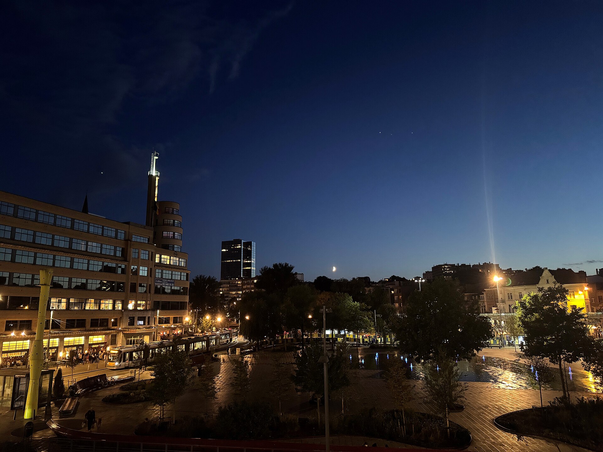 The illuminated Flagey building overlooks Place Flagey in Ixelles at night, with a tram, cafe customers and fountains beneath the moon.