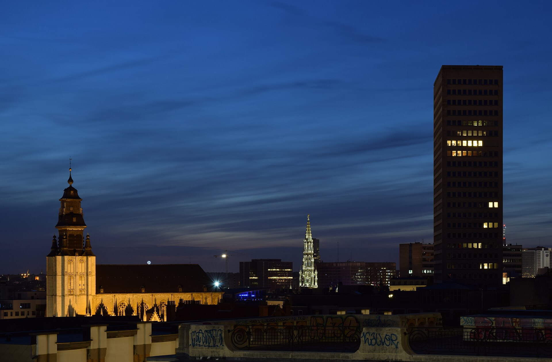 Illuminated Notre-Dame de la Chapelle and the Town Hall spire rise over Brussels rooftops at dusk, seen from the Palais de Justice.
