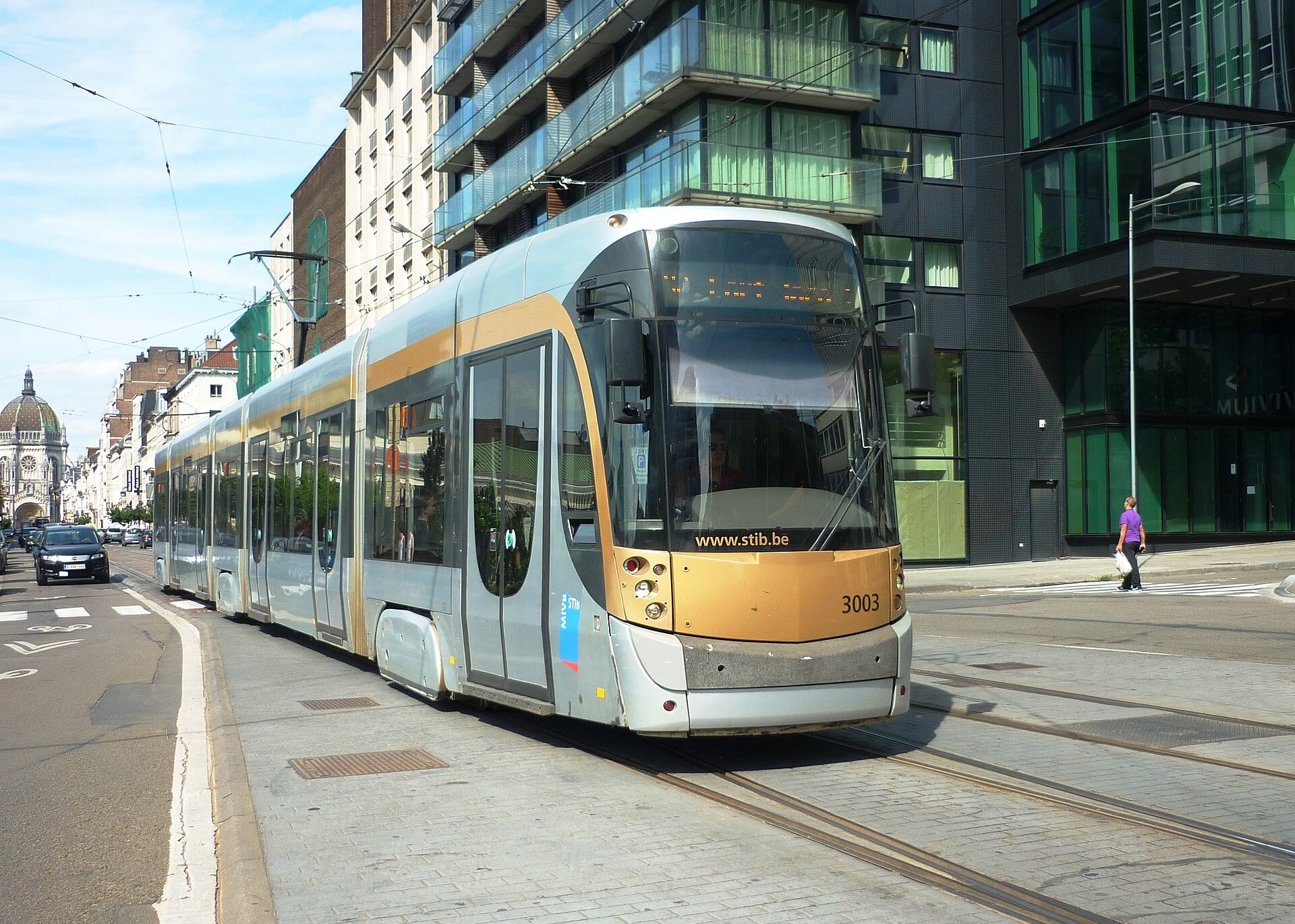 Low-floor STIB tram 3003 travels along a Brussels street, with apartment buildings and a domed church in the background.