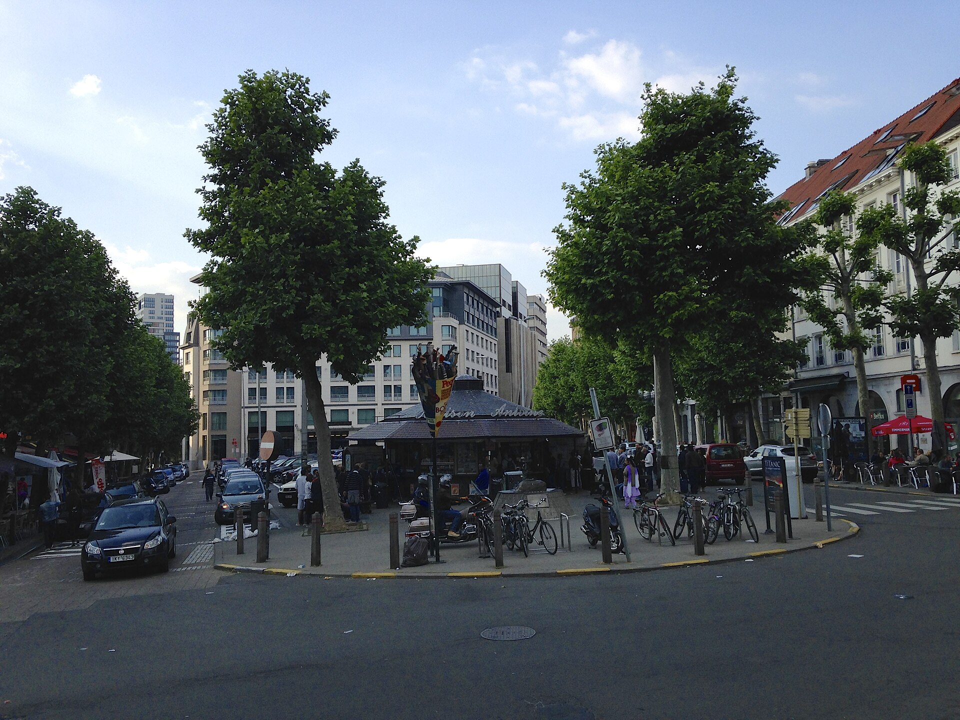 Maison Antoine stands between trees on Place Jourdan in Brussels, with customers, parked bicycles and cafe terraces around the kiosk.