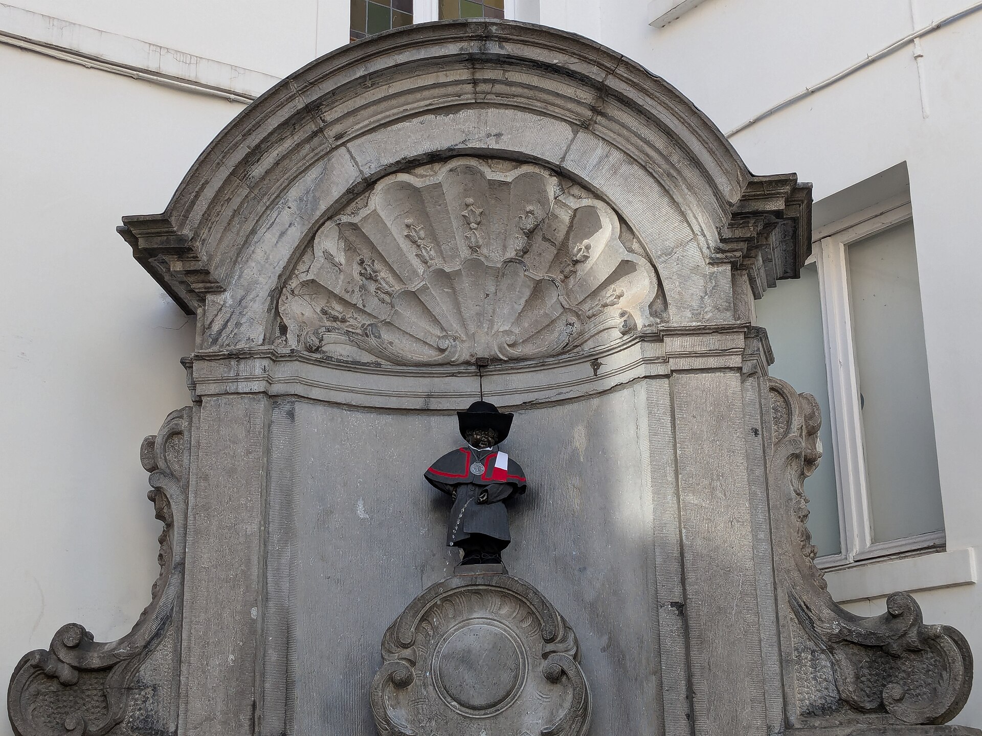 Manneken Pis wears a black hat and red-trimmed dark costume beneath the carved shell of its Brussels fountain niche.
