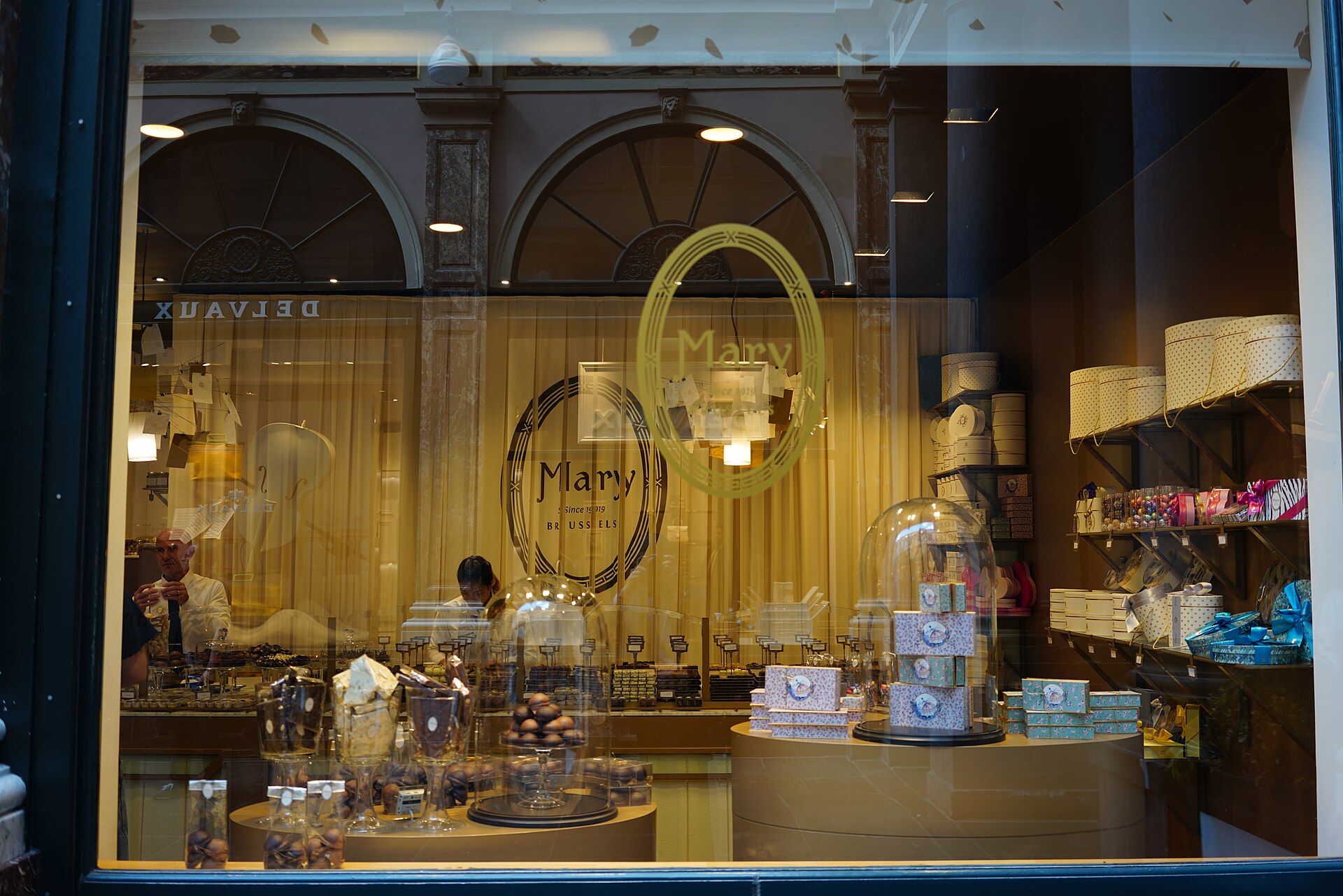 Mary chocolatier in Brussels displays chocolates under glass domes beside pastel gift boxes, with the covered gallery reflected in its window.