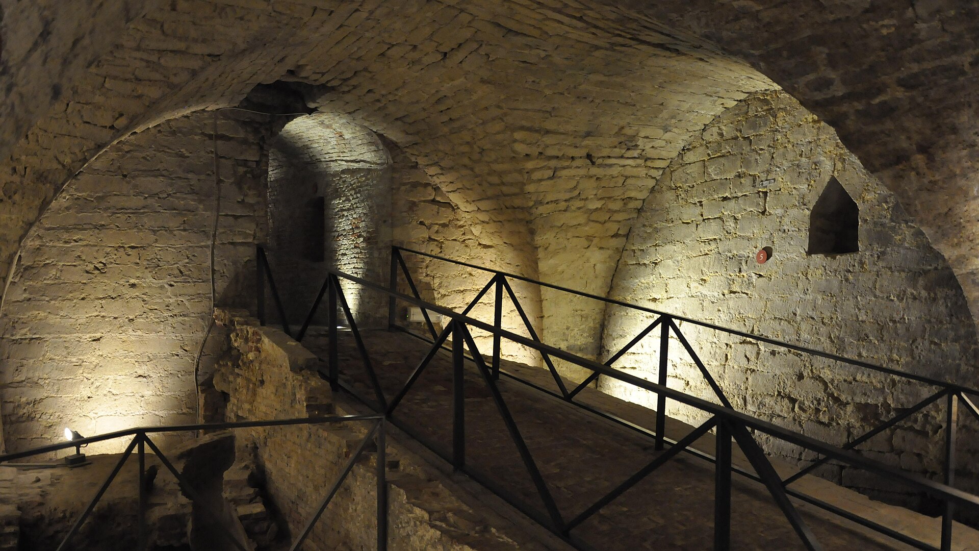A metal-railed walkway passes beneath brick vaults among the illuminated underground remains of Coudenberg Palace in Brussels.