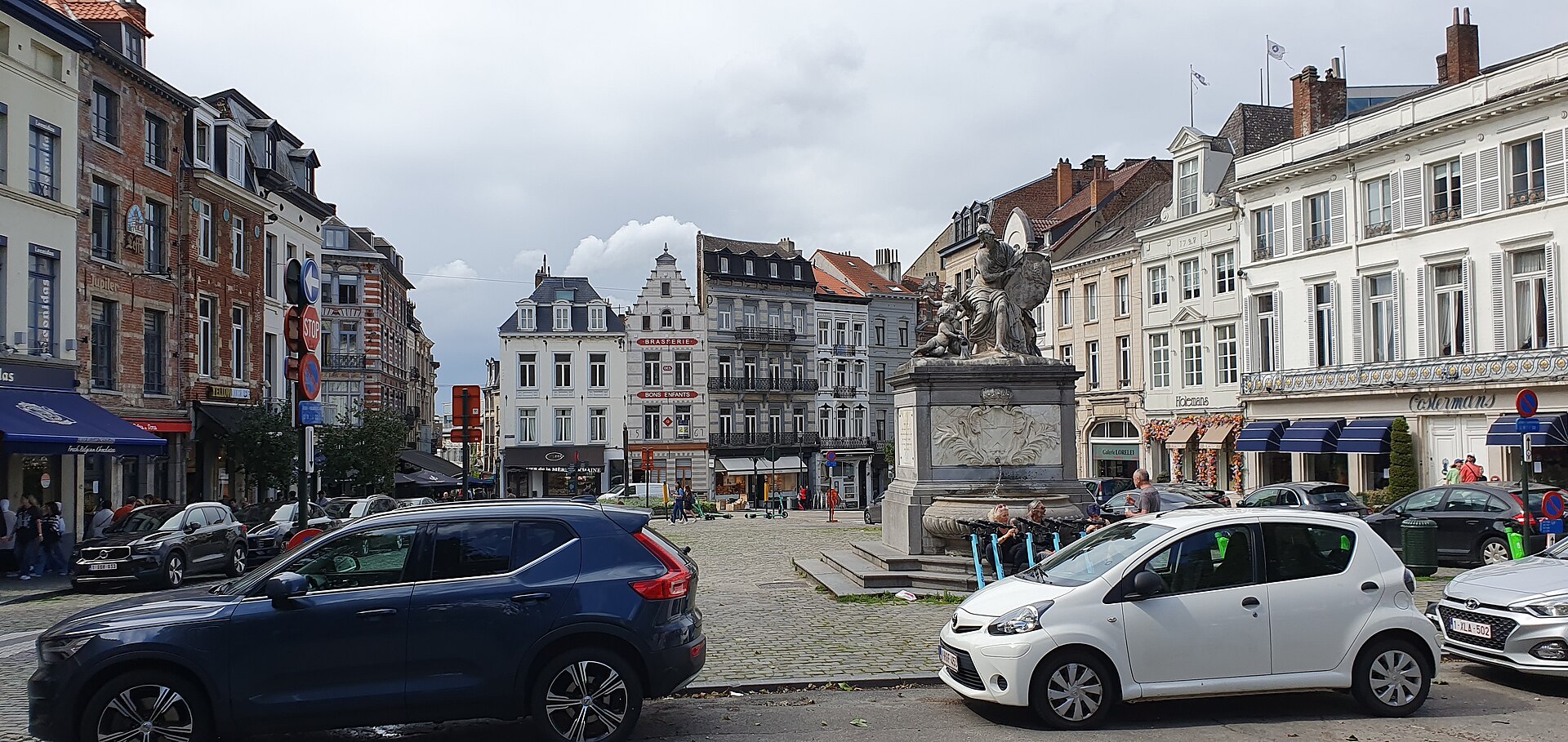 The Minerva Fountain stands among cobbles and historic shopfronts on Place du Grand Sablon in Brussels, with cars passing.