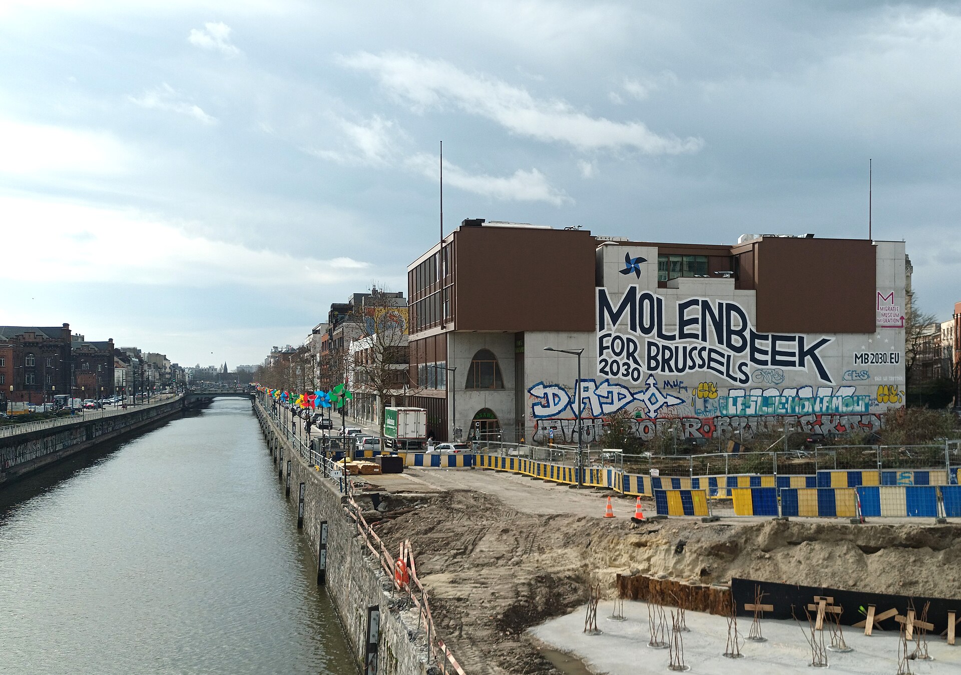 A Molenbeek for Brussels 2030 mural overlooks the canal, with graffiti and waterfront construction beneath a cloudy sky in Brussels.