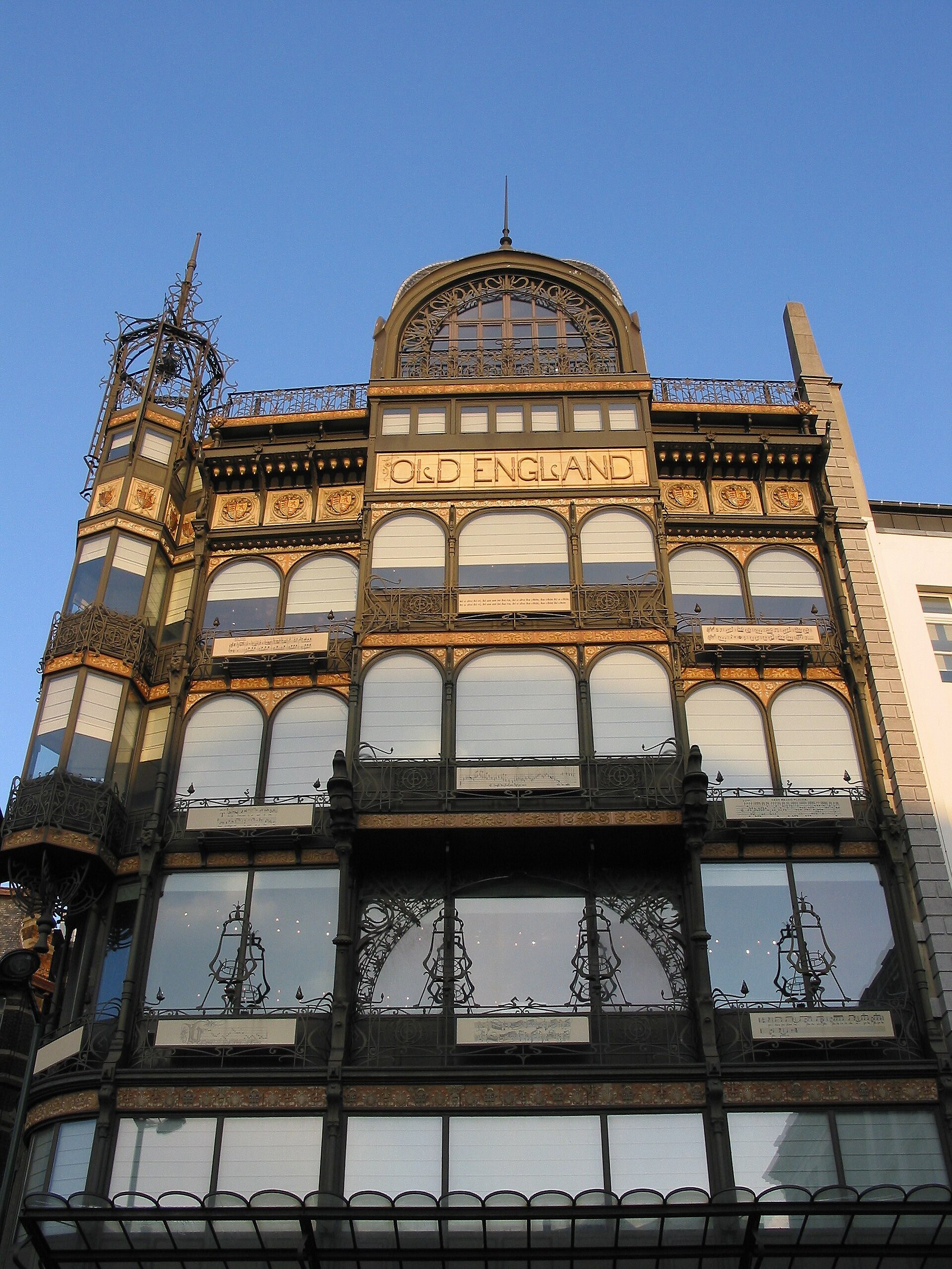The Old England building in Brussels displays ornate iron balconies, tall windows and its original name beneath an arched roof.