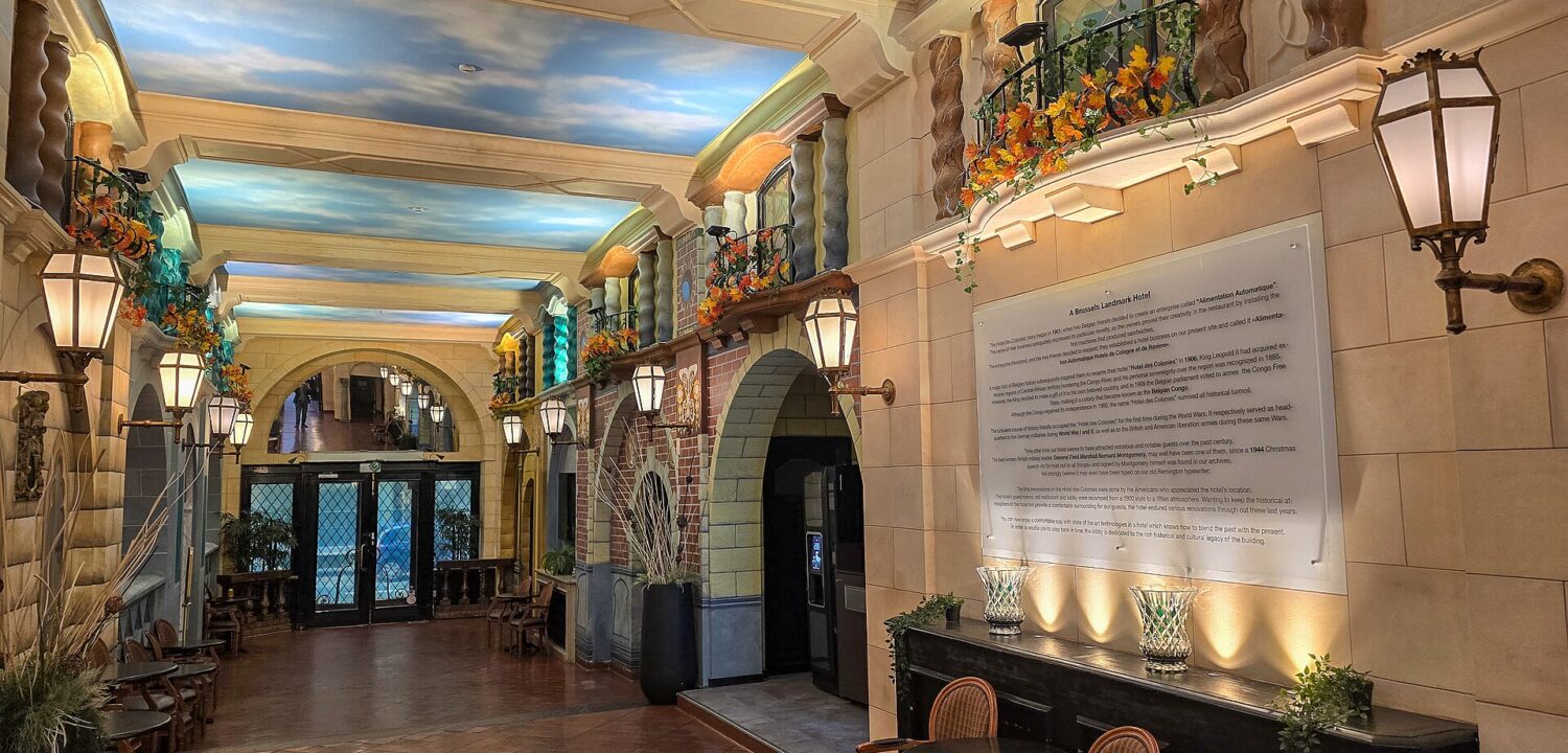 Painted sky ceilings, lanterns and decorative balconies line the Hotel des Colonies lobby in Brussels, with chairs beside a glass doorway.