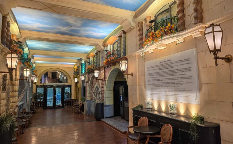Painted sky ceilings, lanterns and decorative balconies line the Hotel des Colonies lobby in Brussels, with chairs beside a glass doorway.