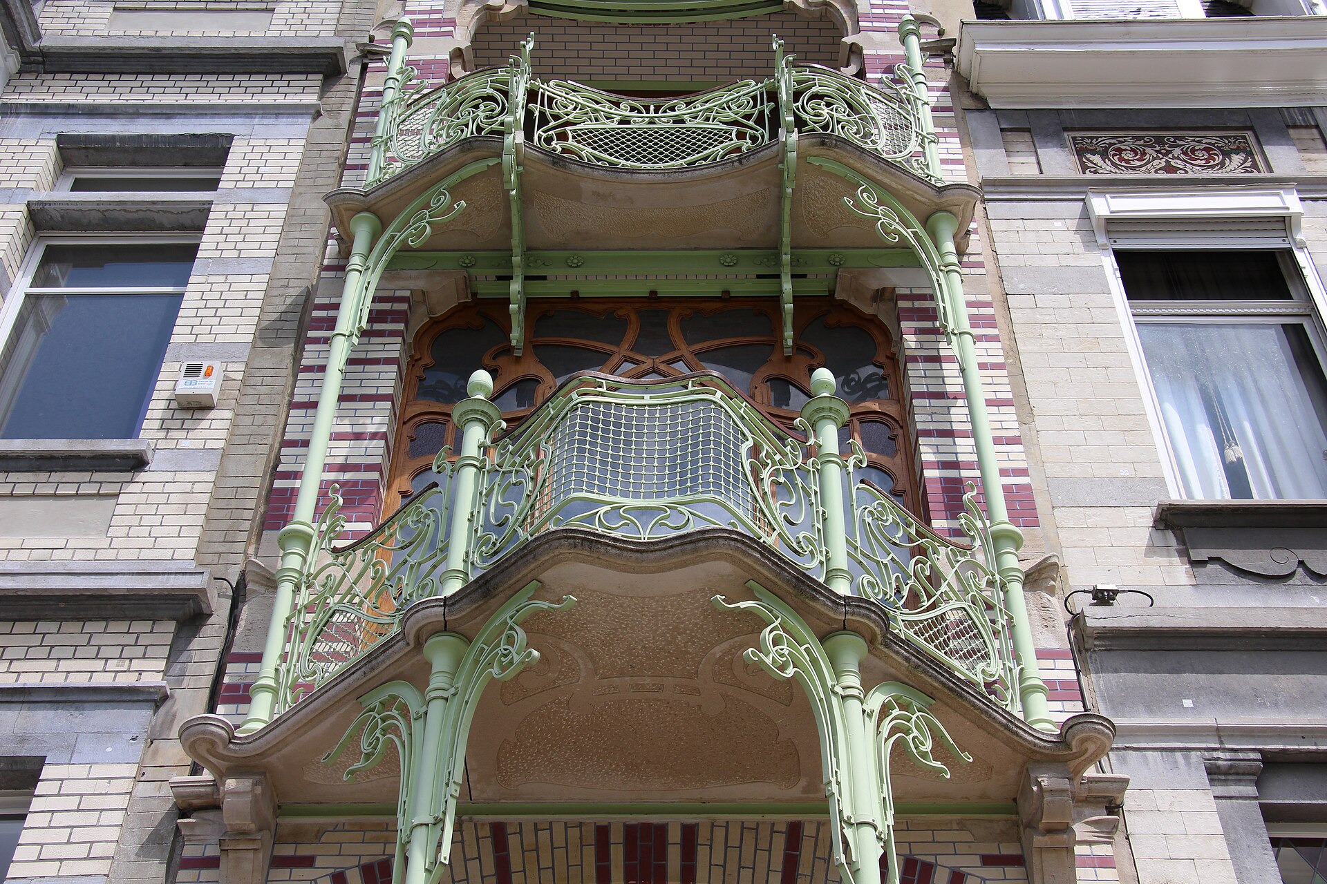 Pale green iron tendrils curl around the balconies of Maison Saint-Cyr in Brussels, above decorative brickwork and wooden windows.