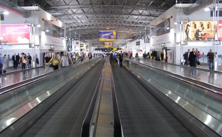 Parallel moving walkways carry travellers through the departures area at Brussels Airport beneath gate signs and a steel roof.