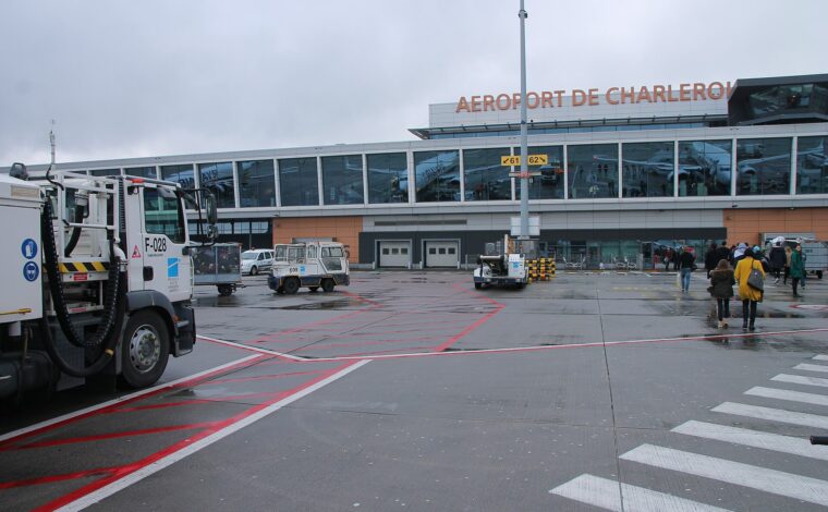 Passengers approach the glass terminal at Charleroi Airport across a wet apron beside ground service vehicles.
