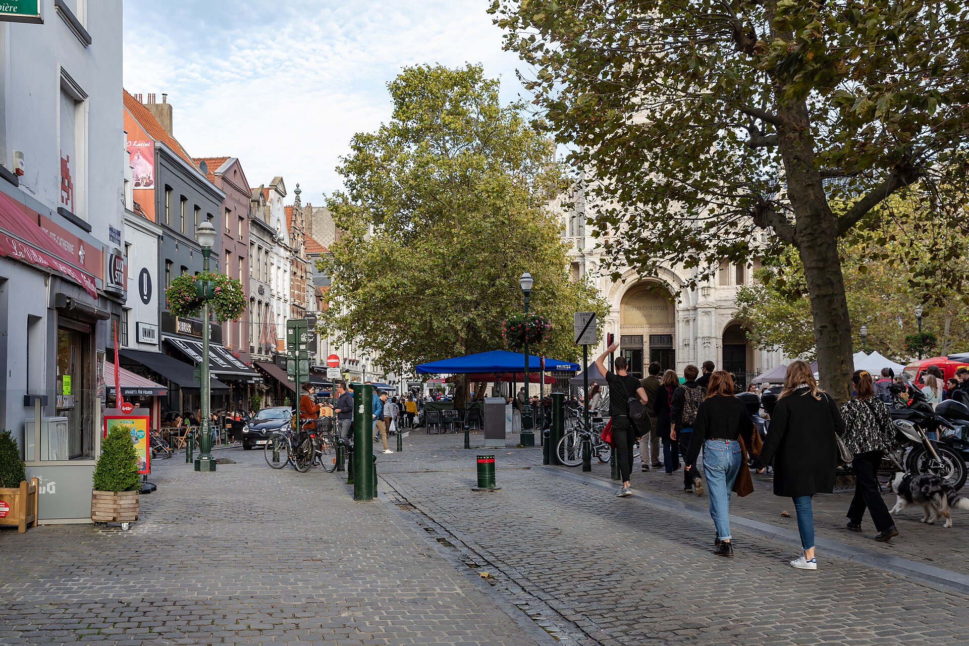 Pedestrians approach Sainte-Catherine church in Brussels along cobbled streets lined with cafes, bicycles and trees on an autumn afternoon.