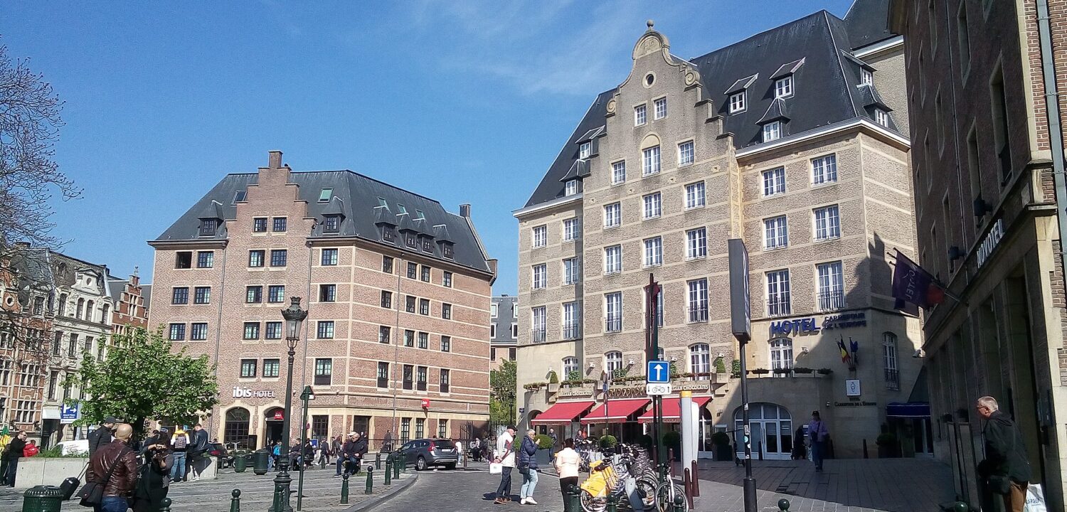 Pedestrians cross a sunny Brussels square between Ibis and Carrefour de l'Europe, with Novotel signage visible at the right.