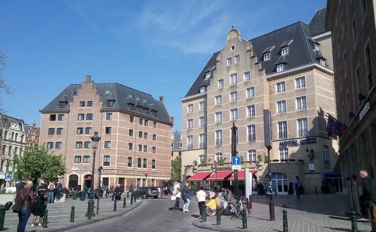 Pedestrians cross a sunny Brussels square between Ibis and Carrefour de l'Europe, with Novotel signage visible at the right.