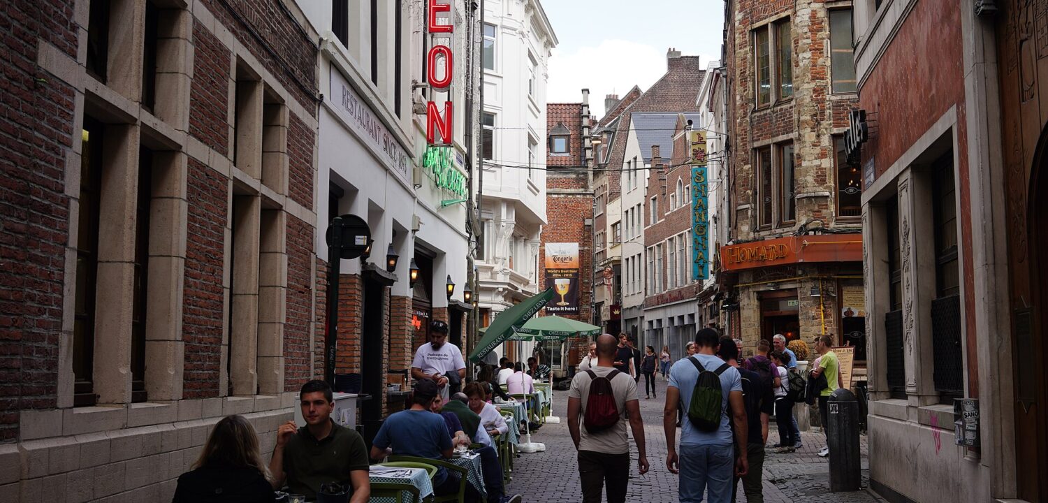 Pedestrians explore Rue des Bouchers in central Brussels between historic brick facades, outdoor restaurant tables and the green umbrellas of Chez Leon.