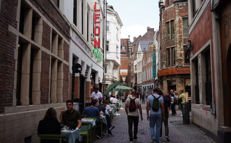 Pedestrians explore Rue des Bouchers in central Brussels between historic brick facades, outdoor restaurant tables and the green umbrellas of Chez Leon.
