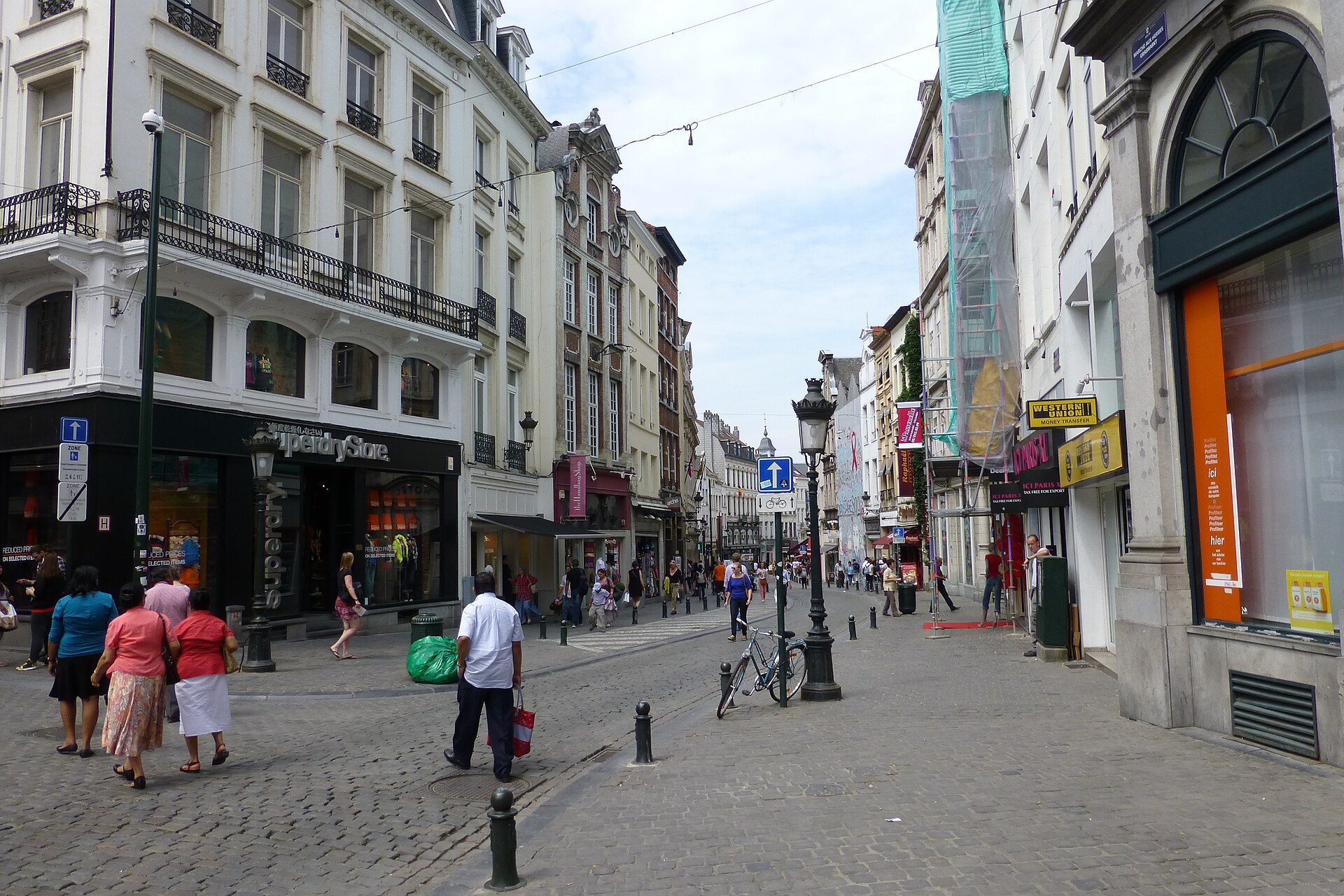 Pedestrians walk along cobbled Rue du Marche aux Herbes in Brussels past historic facades, traditional street lamps and a parked bicycle.